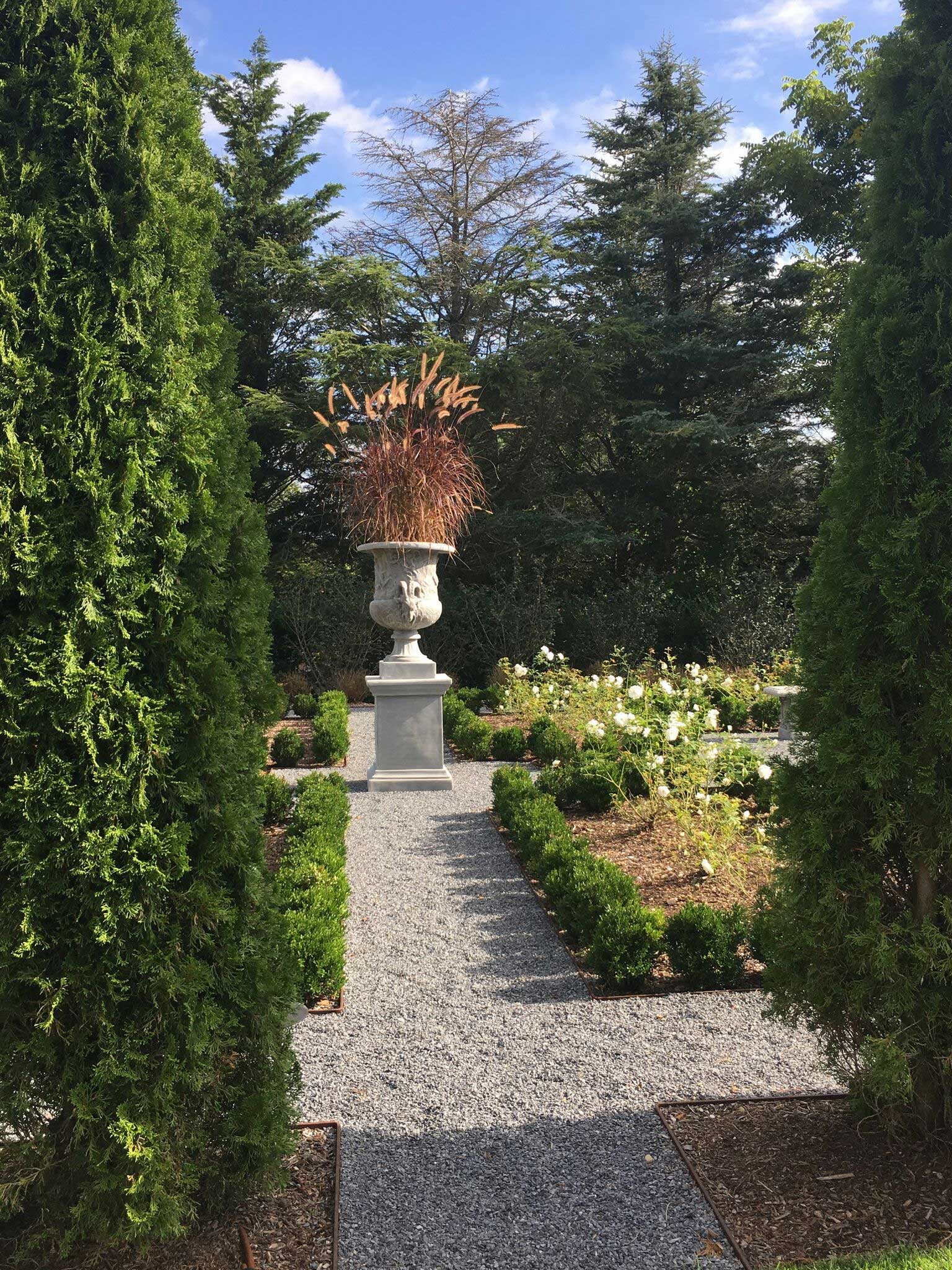 Behind the house, passing through an Arborvitae hedge, one arrives at a Parterre Garden. At the intersection of the gravel pathways on either end of the garden are oversized urns with plantings that rotate through the seasons. The ornamental grasses are coloring up in the early fall.